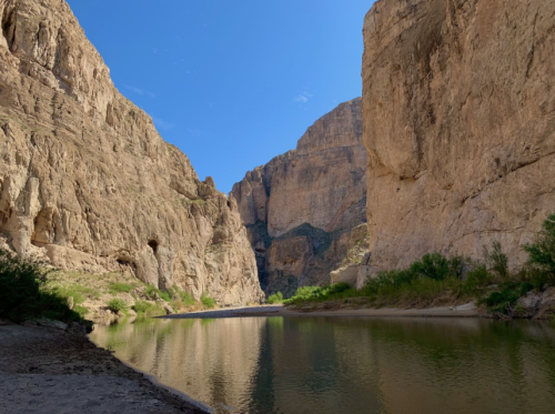 A serene river flows between towering rocky cliffs under a clear blue sky. Lush greenery lines the water's edge.