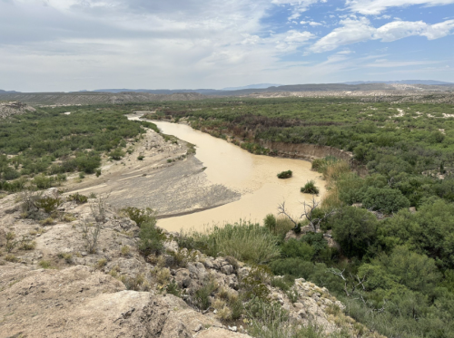 A winding river flows through a green landscape, surrounded by rocky terrain and under a partly cloudy sky.