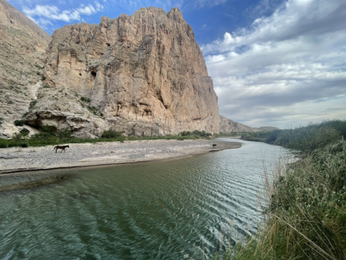 A serene river curves around a rocky cliff, with lush greenery and a horse grazing along the shore under a cloudy sky.