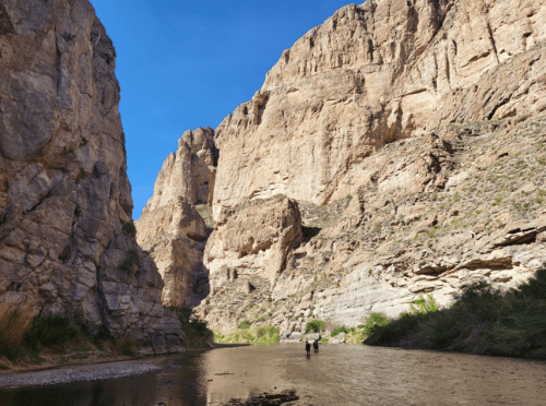Two people walk along a river surrounded by towering rock formations and a clear blue sky.