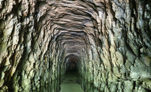 A dimly lit underground tunnel with rocky walls and a shallow waterway running through it.