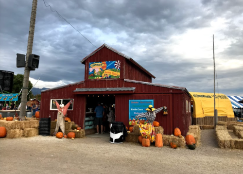 A red barn with pumpkins outside, surrounded by hay bales, under a cloudy sky at a fall festival.