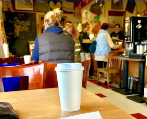 A white coffee cup sits on a table in a busy café with people chatting and ordering in the background.