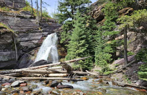 A serene waterfall cascades down rocky cliffs, surrounded by lush green trees and fallen logs in a tranquil forest setting.