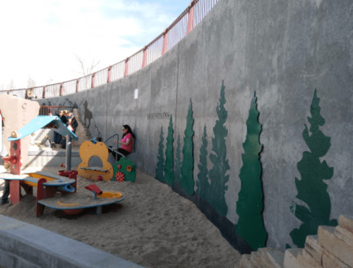 A playground with colorful equipment and a sandy area, surrounded by a curved wall featuring green tree silhouettes.