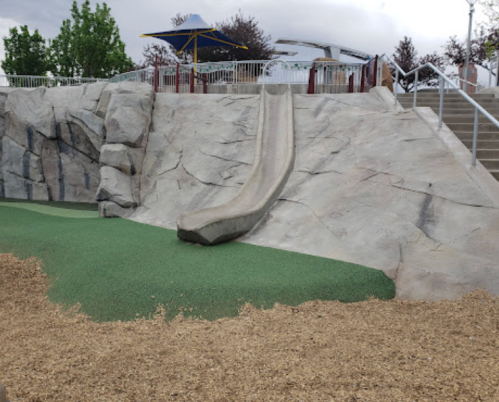A playground slide built into a rocky structure, surrounded by green turf and wood chips, with trees in the background.