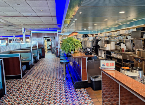 A brightly lit diner interior featuring booths, a counter, and a kitchen area with a colorful tiled floor.