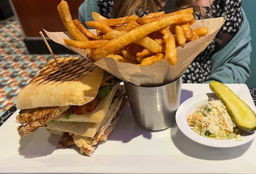 A plate with a grilled sandwich, a side of crispy fries in a cup, coleslaw, and a pickle slice.