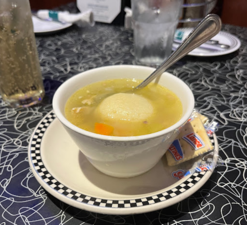 A bowl of soup with a dumpling, served on a plate with crackers, on a patterned tablecloth.