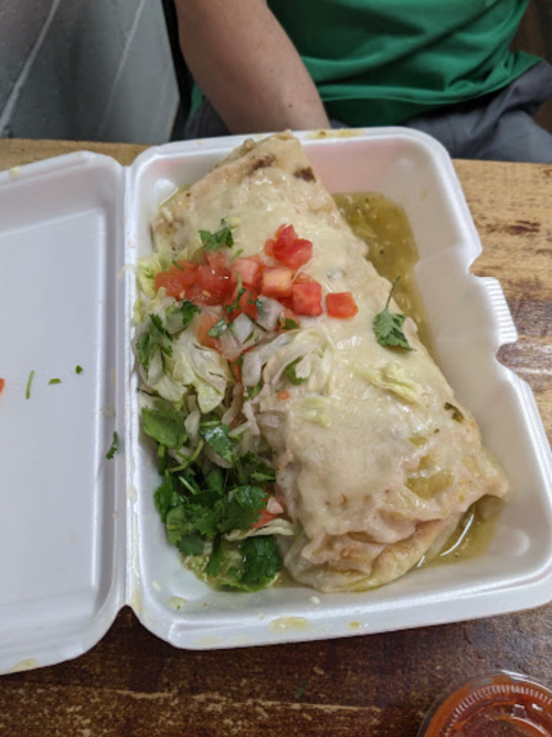 A large burrito topped with cheese, tomatoes, and lettuce, served in a takeout container with green sauce on the side.