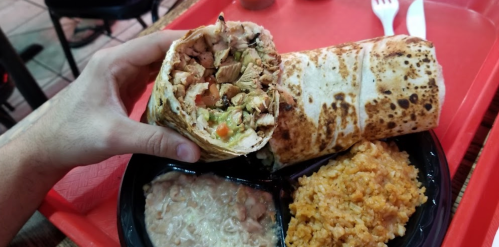 A hand holding a chicken burrito next to rice and beans on a red tray.