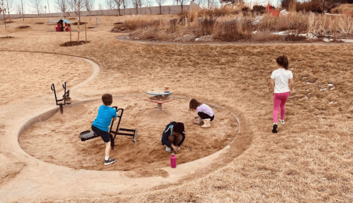 Children playing in a sandy area at a park, with some digging and others walking nearby.