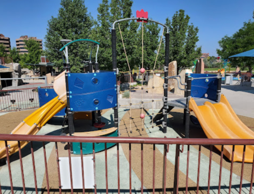 Colorful playground with slides, climbing structures, and swings, surrounded by trees and a clear blue sky.