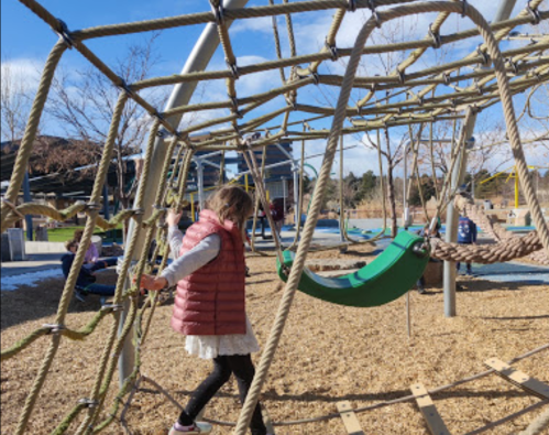 A child in a pink vest climbs a rope structure at a playground with swings and trees in the background.