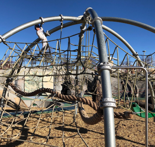 A playground structure with climbing ropes and a clear blue sky in the background. A child is climbing on top.
