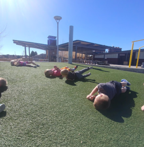 Children playing on a grassy field, lying on their stomachs under a clear blue sky.