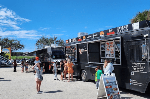 A busy food truck event with people lining up at a black food truck under a clear blue sky.