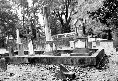 Black and white image of a cemetery with various gravestones and a surrounding fence, set among fallen leaves.