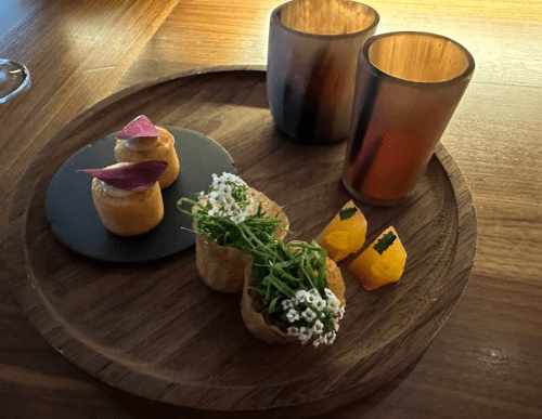 A wooden platter with two colorful glasses, gourmet appetizers, and edible flowers arranged artistically.