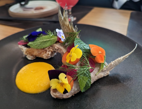 A beautifully plated dish featuring fried fish garnished with edible flowers and herbs, served with a yellow sauce.