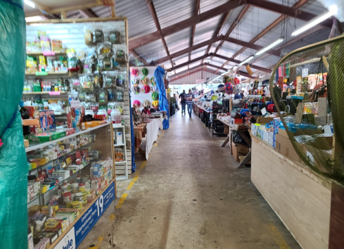 A bustling market aisle lined with various goods, including crafts, toys, and household items, under a metal roof.