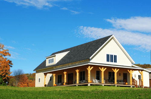 A yellow house with a porch and wooden pillars, set against a blue sky and green lawn, surrounded by autumn trees.