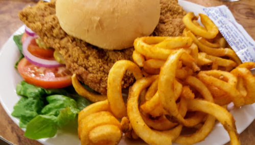 A plate featuring a fried chicken sandwich on a bun, surrounded by lettuce, tomato, onion, and curly fries.