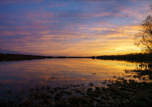 A serene lake at sunset, reflecting colorful clouds and trees along the shore.