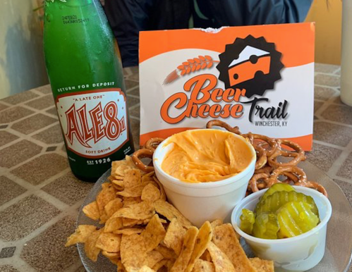 A plate with cheese dip, pretzels, chips, and pickles, alongside a bottle of Ale-8 soda and a Beer Cheese Trail sign.