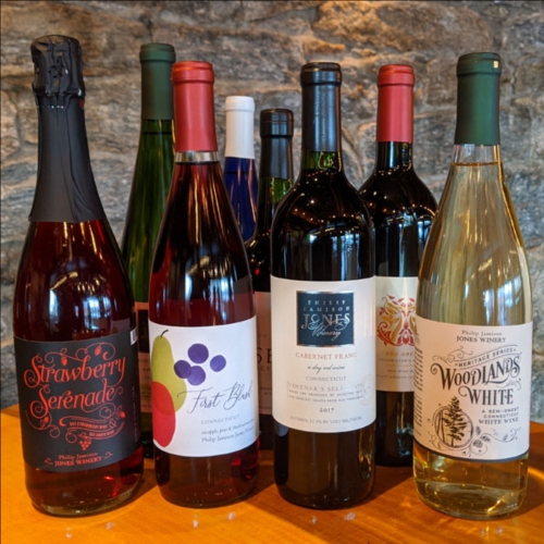 A selection of wine bottles, including sparkling, red, and white varieties, displayed on a wooden table against a stone wall.