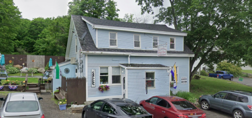 A light blue two-story building with a sign, surrounded by trees and parked cars in a small lot.