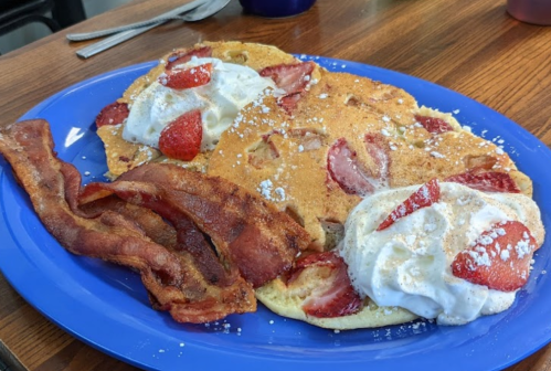 A blue plate with two strawberry pancakes topped with whipped cream and bacon strips on the side.