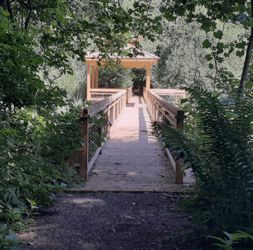 A wooden bridge leads to a gazebo, surrounded by lush greenery and trees.