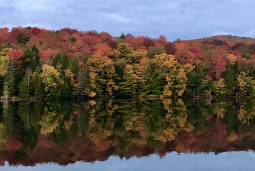 Vibrant autumn foliage reflects on a calm lake, showcasing shades of red, orange, and yellow against a serene sky.