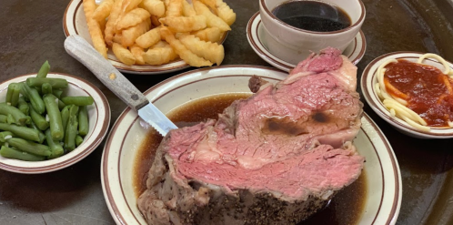 A plate with a large slice of prime rib, green beans, crinkle fries, and spaghetti with sauce, served with gravy.