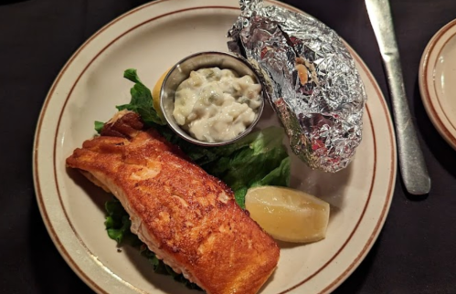 A plate with grilled salmon on lettuce, a baked potato wrapped in foil, and a small bowl of sauce, garnished with lemon.