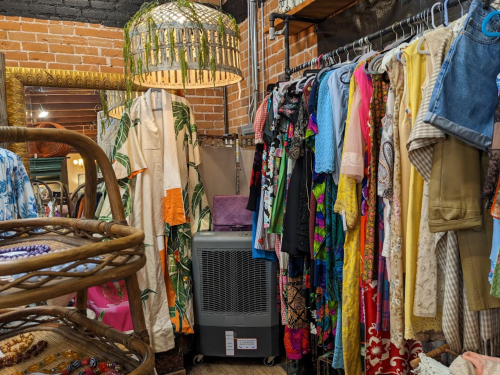 A vintage clothing rack filled with colorful garments, next to a large fan and a decorative hanging light fixture.
