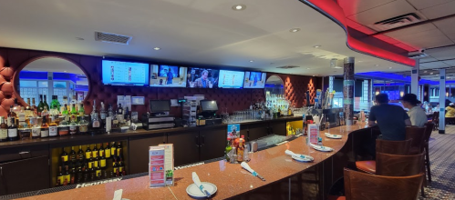 A modern bar with a well-stocked counter, two patrons seated, and multiple screens displaying drinks and entertainment.