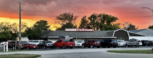 A steakhouse at sunset, with a vibrant orange sky and a parking lot filled with cars.