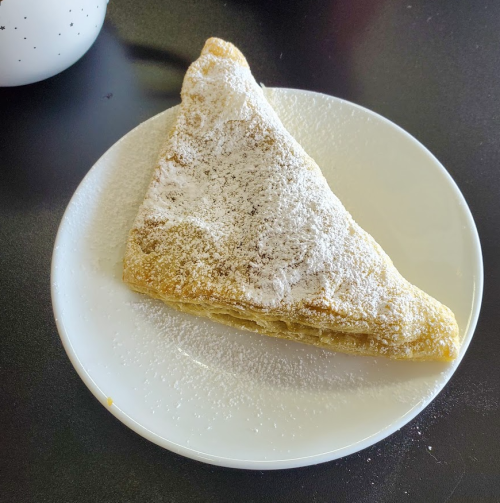 A triangular pastry dusted with powdered sugar on a white plate, resting on a dark surface.