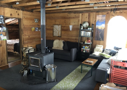 Cozy wooden living room with a wood stove, two sofas, a coffee table, and a bookshelf, featuring warm, rustic decor.