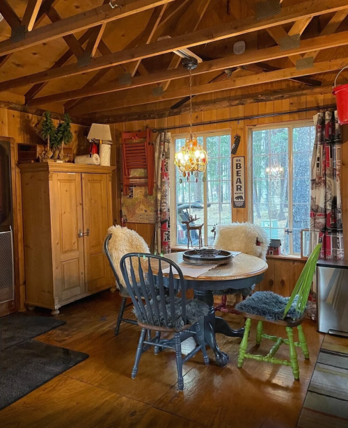 Cozy cabin interior with a wooden table, chairs, and large windows, surrounded by rustic decor and warm lighting.