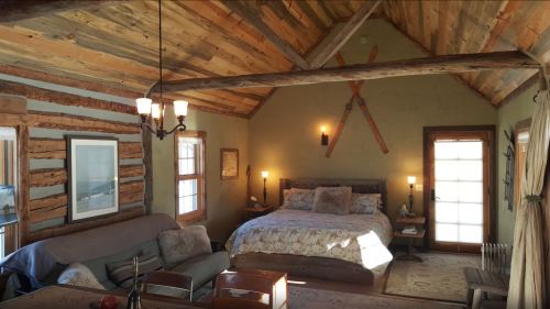 Cozy cabin interior featuring a bed, sofa, wooden beams, and rustic decor with natural light from windows.