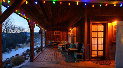 Cozy porch with colorful lights, wooden walls, and snow-covered landscape at dusk.