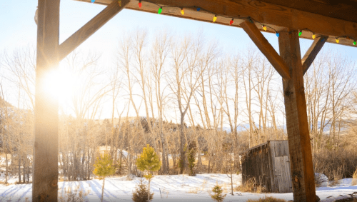 Sunlight filters through trees in a snowy landscape, framed by a wooden porch with colorful holiday lights.