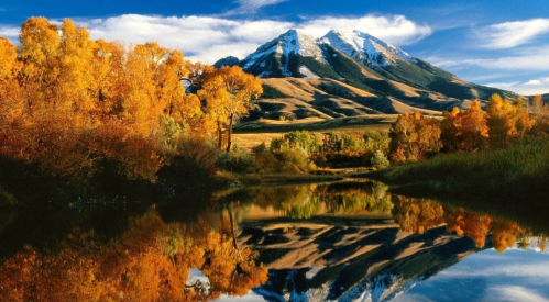 A serene landscape featuring autumn trees, a reflective lake, and snow-capped mountains under a blue sky.