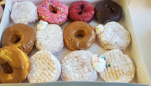 A box of assorted donuts, including glazed, powdered, and frosted varieties, with colorful sprinkles and cream toppings.