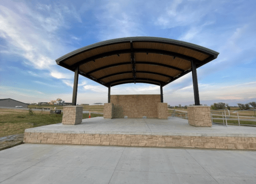 A large outdoor stage with a curved roof, set against a clear sky and grassy area.