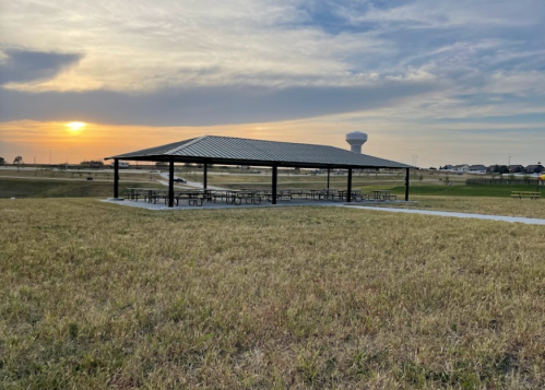 A metal pavilion with picnic tables at sunset, surrounded by grassy fields and a water tower in the background.