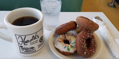 A cup of coffee next to a plate of assorted donuts with sprinkles and a glass of water on a table.